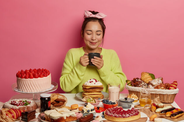 煎饼亚洲女人戴着头巾 喝茶 围着美味的甜点 拿着杯子 闭着眼睛 隔离在粉红色的墙上甜食爱好者享受美味的早餐草莓甜的营养