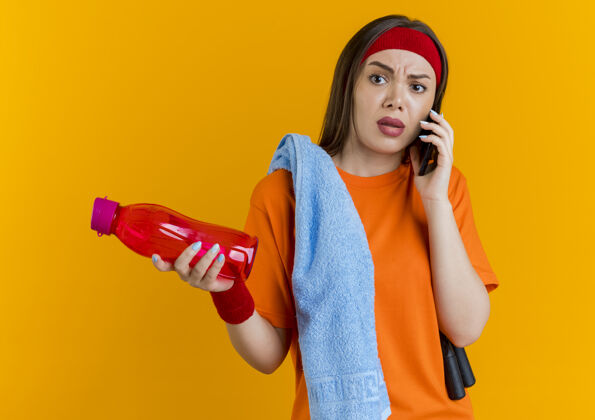 橙色戴着头带和腕带 肩上扛着跳绳和毛巾 手里拿着水瓶 边打电话边聊天的年轻运动型女人穿毛巾绳子