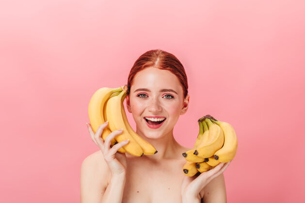 香蕉令人惊叹的姜汁女孩与香蕉的正面视图摄影棚拍摄的快乐裸体女子手持粉红色背景上的热带水果微笑裸体女人