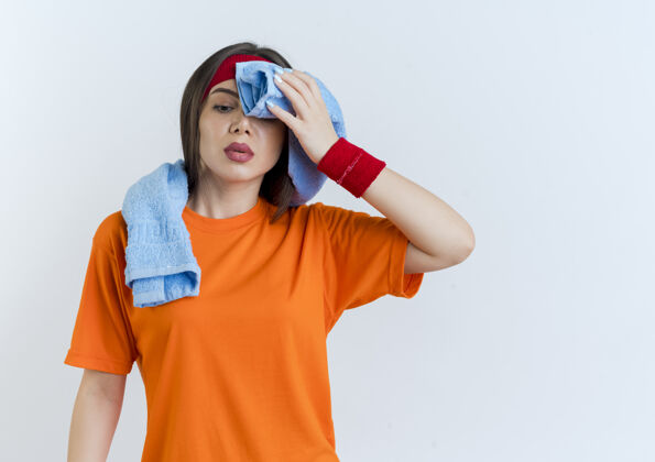 腕带疲惫的年轻运动女性戴着头带和手环 脖子上围着毛巾 低头用毛巾擦汗运动累了汗