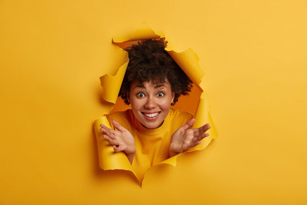感觉快乐的非裔美国女人穿着黄色毛衣 摊开手掌 露出洁白的牙齿 在室内玩得很开心 透过撕破的纸洞看背景快乐洞情绪