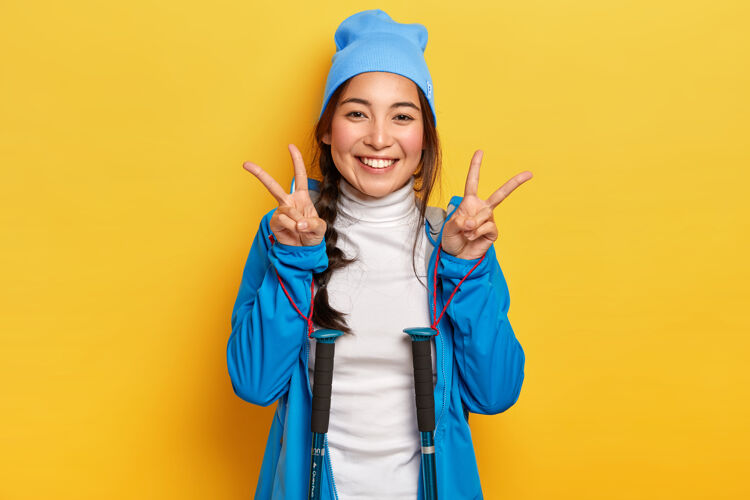 人快乐的女人摆出和平的姿态 拿着登山竿摆姿势 戴着蓝色的帽子和夹克 喜欢徒步旅行 高兴地看着镜头 隔着黄色的墙活动高兴徒步旅行