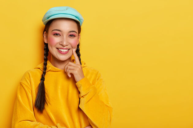 站迷人的亚洲女士 粉色妆容 食指贴在脸颊上 思考着好主意 穿着休闲的黄色丝绒运动衫 带着兜帽 微笑温柔辫子帽子头巾