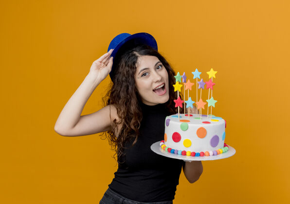 卷发一个年轻漂亮的女人 卷发 戴着节日帽 手里拿着生日蛋糕 快乐地站在橘色的墙上女人站着看着