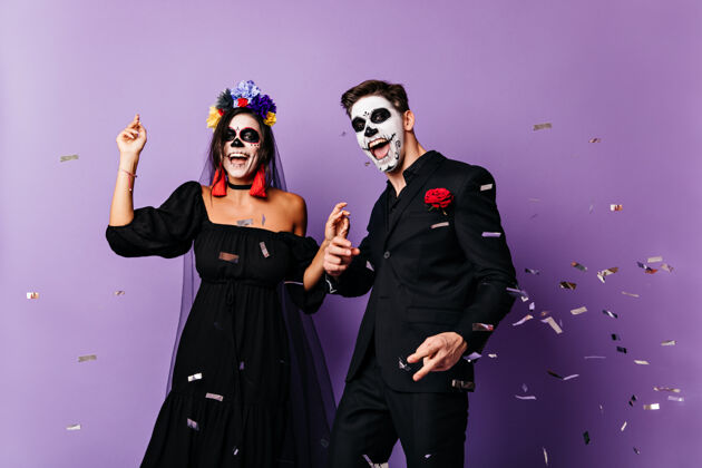 死亡之日有趣的家伙和黑头发的女士画了脸和花冠摆姿势 穿着黑色服装跳舞的聚会香槟男人骷髅