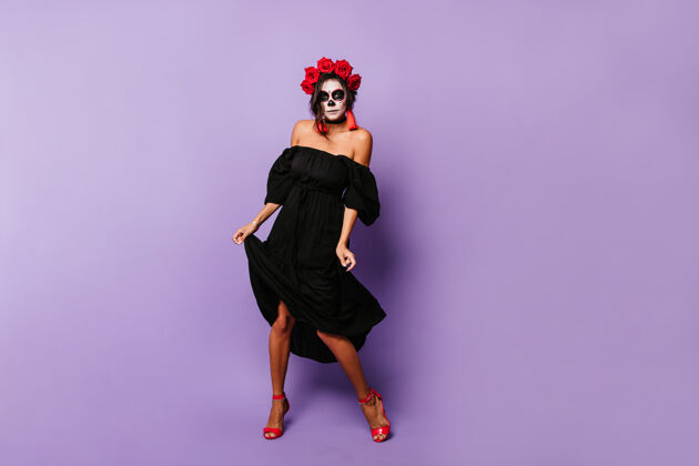 花活跃的墨西哥骷髅女郎在淡紫色的墙上翩翩起舞身着红色配饰和玫瑰的女士摆出全身照女人民间传说人体艺术