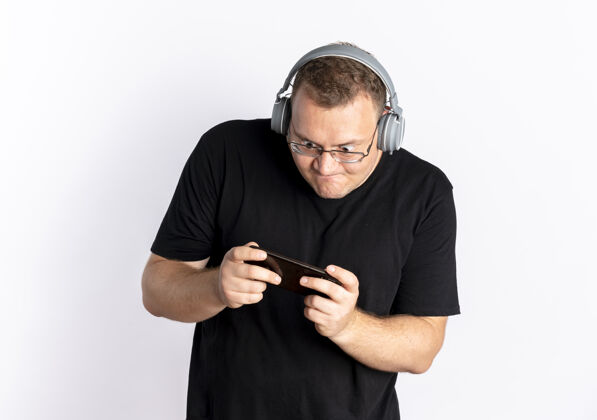 玩一个戴眼镜的超重男人 穿着黑色t恤 戴着耳机 站在白墙上用智能手机玩游戏眼镜耳机手机