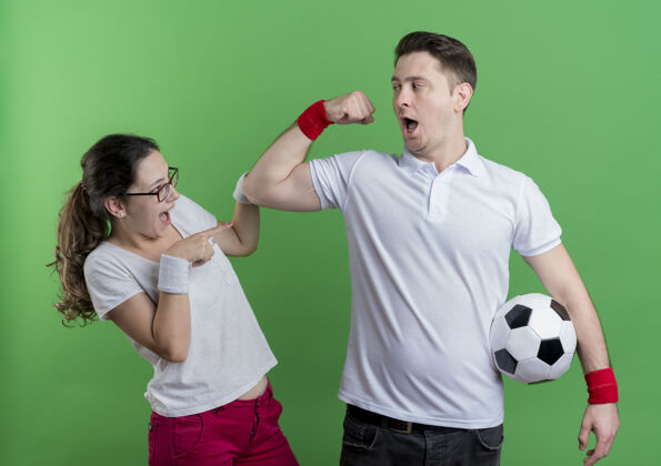 足球一对年轻的运动型情侣站在绿色的墙上 拿着足球向他惊讶的女友展示二头肌站情侣表演