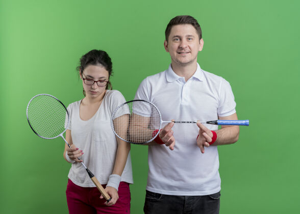 网球一对年轻的运动情侣手拿球拍打网球 微笑着站在绿色的墙上情侣运动微笑