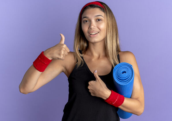 年轻身穿黑色运动服 头戴红色头巾的年轻健身女孩手持瑜伽垫 微笑着站在蓝色墙壁上竖起大拇指看着镜头运动装看瑜伽