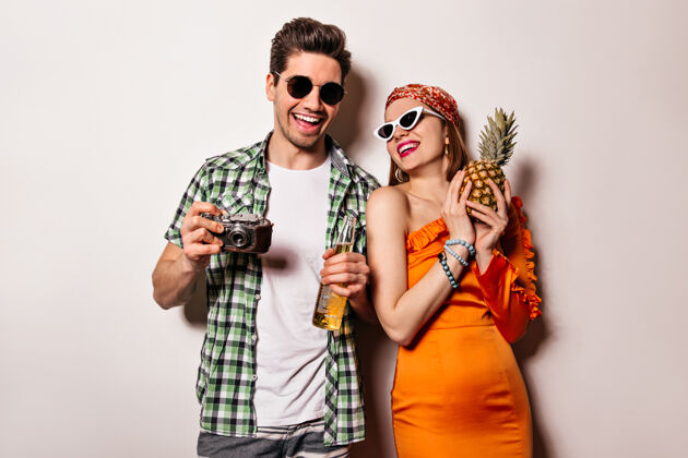 衬衫戴着墨镜 穿着时髦的橙色套装的迷人女孩和她的男朋友在白色空间里摆姿势 手里拿着菠萝 复古相机和一瓶啤酒手菠萝橙色连衣裙