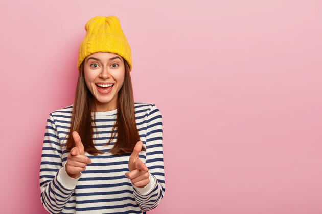 复制空间迷人的年轻女性穆德尔的照片做手指势 戴黄色帽子和条纹套头衫 表达选择 选择你到她的团队 孤立在粉红色背景上显示套头衫微笑