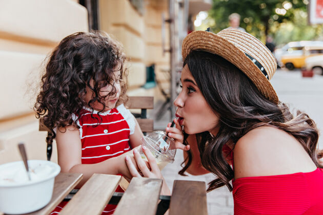 情绪黑发女人戴着草帽和女儿在咖啡馆里玩可爱的小女孩坐在户外餐厅看着妈妈妈妈冰淇淋户外