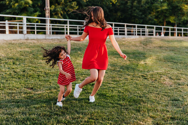 女人一位身着红色短裙的优雅小姐与女儿手牵手母亲和孩子在公园玩耍的户外全幅照片妈妈草地年轻