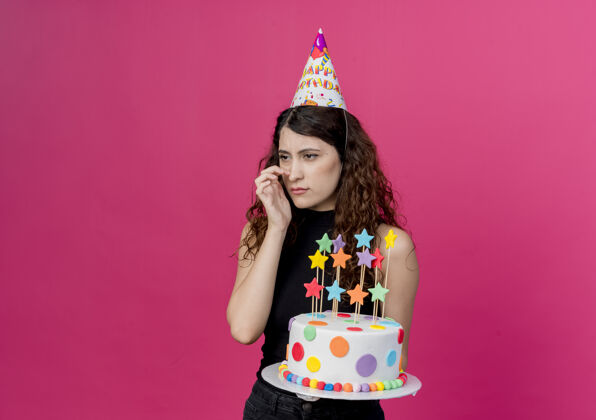 抱着一个年轻漂亮的女人 卷发 戴着节日帽 手里拿着生日蛋糕 站在粉色的墙上 一边看着困惑的生日派对概念站在一边看女人