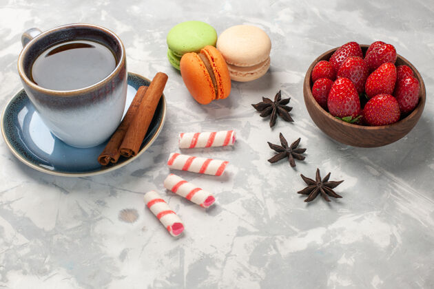 视图正面图法式麦卡龙配上一杯茶和新鲜的红色草莓在白色表面蛋糕糖饼干甜饼干蛋糕麦卡龙杯子