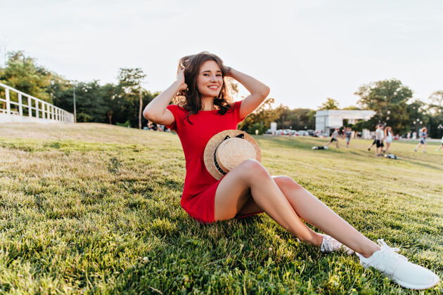 和平积极的白人女孩坐在草地上 玩着她的棕色头发快乐的笑着的红衣女士在公园享受周末的早晨田野时尚女人