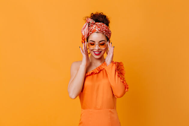 服装戴着橙色眼镜 丝质连衣裙和头带的可爱女人在橙色的空间里微笑着长橙色连衣裙蓝眼睛