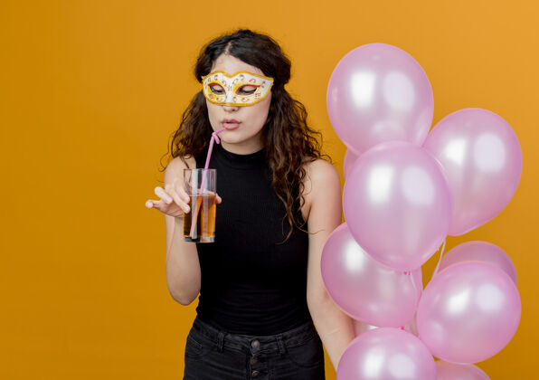 站着年轻漂亮的卷发女子手持一束气球 戴着派对面具 快乐快乐地喝着鸡尾酒 站在橙色的墙上 生日派对的概念举行年轻束