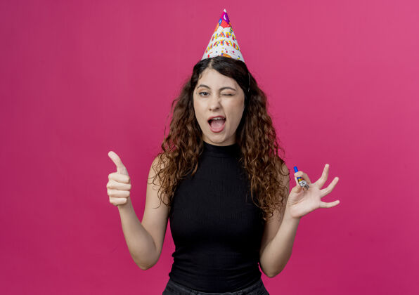 女人一位年轻漂亮的女士 一头卷发 戴着节日帽 微笑着眨眼 竖起大拇指 站在粉色的墙上 庆祝生日派对展示拇指帽子