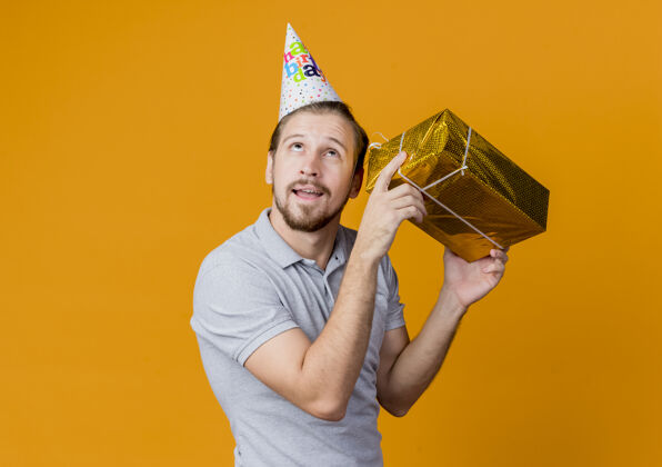 男人戴着节日帽的年轻人举着礼品盒站在橙色的墙上 耳边挂着沉思的表情 庆祝生日聚会表情站立沉思