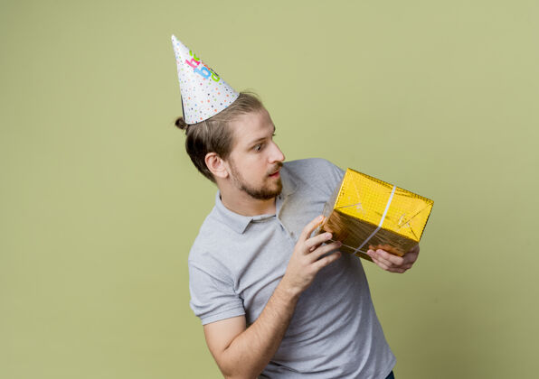 年轻戴着节日帽的年轻人拿着生日礼物看着它惊讶地站在轻墙上的生日派对概念抱着光看着