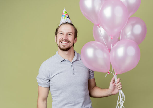 微笑戴着节日帽的年轻人举着气球疯狂地欢庆生日 兴奋地微笑着站在轻墙上庆祝光站立