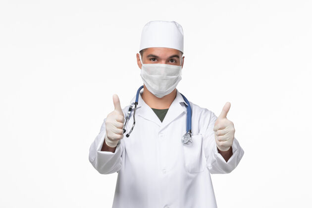 对抗正面图：男医生穿着医疗服 戴着口罩 戴着听诊器 对着白墙病毒-病毒-疾病大流行摆姿势冠状病毒大流行视图