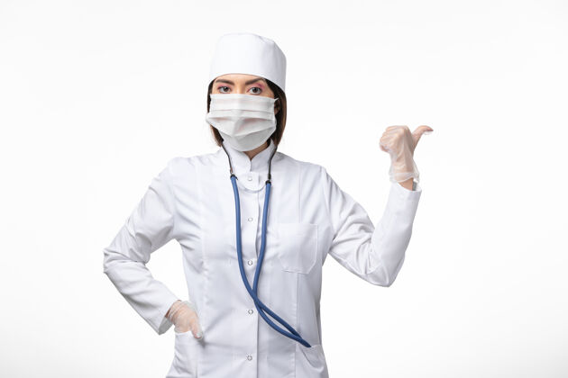大流行前视图女医生穿着白色医疗服 戴着口罩 因大流行摆在白墙上疾病医学病毒大流行冠状病毒-女性医生姿势