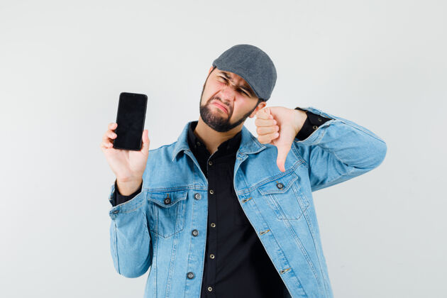 时尚复古风格的男人展示拇指向下 而显示手机在夹克 帽子 衬衫和看起来不满意前视图年轻成人不满意
