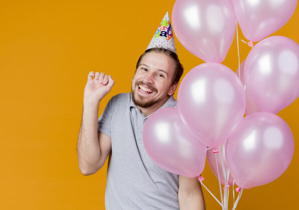 兴奋戴着节日帽的年轻人举着一堆气球 站在橘色的墙上 高兴而兴奋地微笑着庆祝生日欢呼庆祝站立
