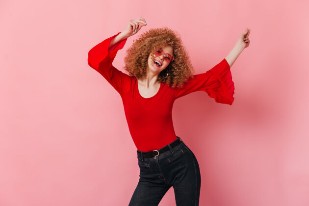 优雅活跃的卷发女士戴着太阳镜 穿着红色长袖上衣和牛仔裤 在粉色的空间里欢笑跳舞人体衬衫魅力