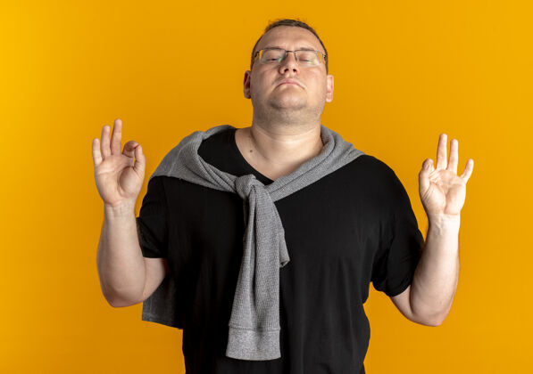 冥想戴眼镜的超重男子穿着黑色t恤 闭着眼睛放松 手指站在橙色的墙上做冥想姿势穿手指站着