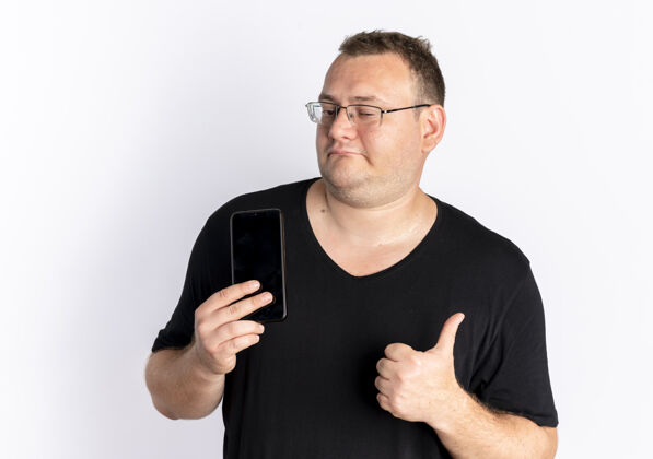 展示戴眼镜的超重男子身穿黑色t恤 手持智能手机 竖起大拇指站在白墙上手机拇指眼镜