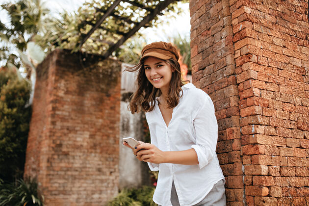 植物可爱的棕色眼睛的女孩微笑着在砖房旁摆姿势戴着帽子和白衬衫的女人拿着智能手机灌木现代温暖