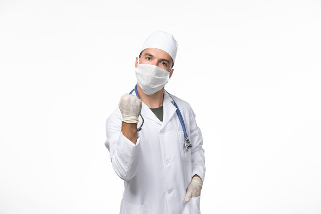冠状病毒正面图：男医生穿着医疗服 戴着防冠状病毒口罩 白色墙壁上有蓝色听诊器 冠状病毒-疾病大流行病毒正面医疗专业