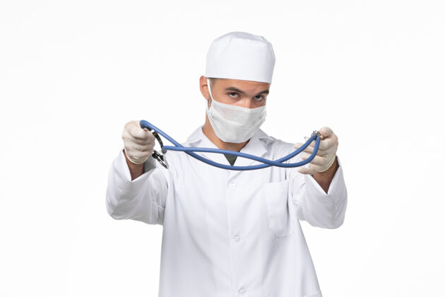 专业正面图：男医生穿着医疗服 戴着口罩 因为白色墙壁上戴着柯维德听诊器 柯维德大流行病毒病持有套装大流行