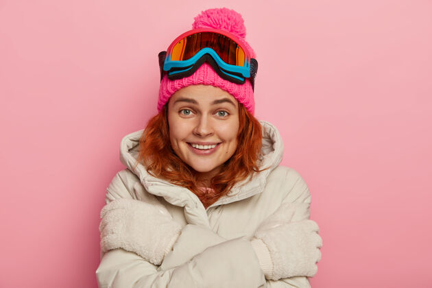 面具活泼好动的女人 表情愉快 红色波浪发 拥抱自己 戴着粉红色的帽子和温暖的外套 隔离在粉红色的背景下姜黄色微笑拥抱