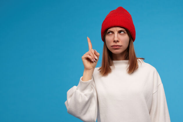 成人漂亮的女人 漂亮的女孩 深色头发穿着白色毛衣 戴着红色帽子抬起手指 有了一个想法看着左边的复制空间 隔着蓝色的墙黑发独处帽子