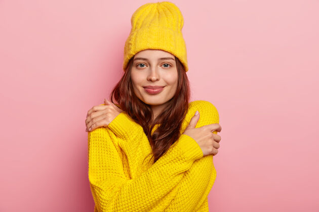 爱横向拍摄的迷人的年轻女子拥抱自己 深色长发 温柔的样子 戴着黄色的冬季帽子和毛衣 在粉红色的工作室背景下摆姿势满意温暖超大