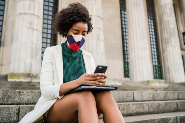 看非洲裔女商人戴着防护面具 坐在街上的楼梯上用手机商业和城市概念冠状病毒户外短信