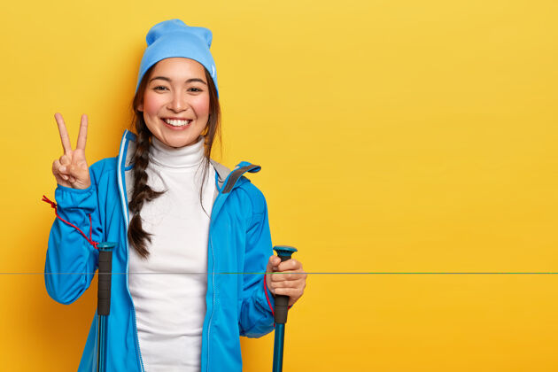 高兴美丽的韩国女孩喜欢徒步旅行 摆姿势与徒步杖 使和平的姿态 戴着蓝色帽子和夹克 隔离在黄色背景 空白亚洲人复制空间手势