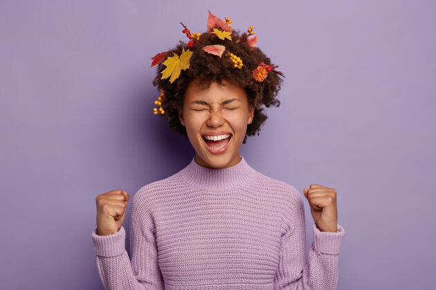 肖像喜出望外的非洲年轻女子的肖像成功地握紧拳头 感到兴奋 有创意的发型装饰着秋天的树叶 穿着暖和的套头衫 孤立在紫色的背景之上欢呼胜利女人