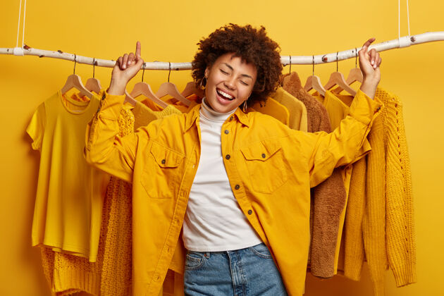 悬挂快乐快乐的非洲女人在衣架上翩翩起舞 喜欢黄色的服装 穿着时髦的夹克和牛仔裤 在衣柜附近积极活动单色欢乐时尚