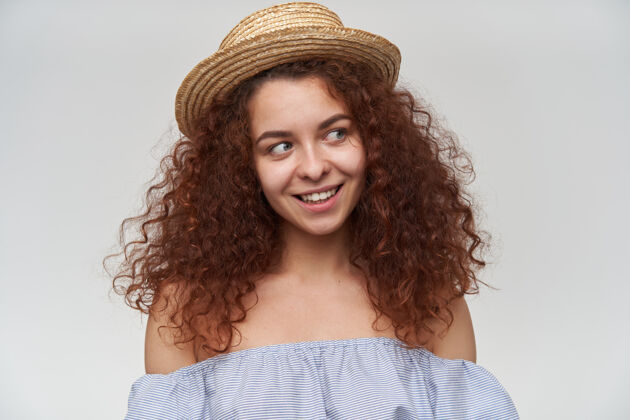 微笑有吸引力的 卷发的成年红发女孩的肖像穿着条纹露肩衬衫和帽子在复制空间看右边 特写 隔着白墙孤立特写女性服装