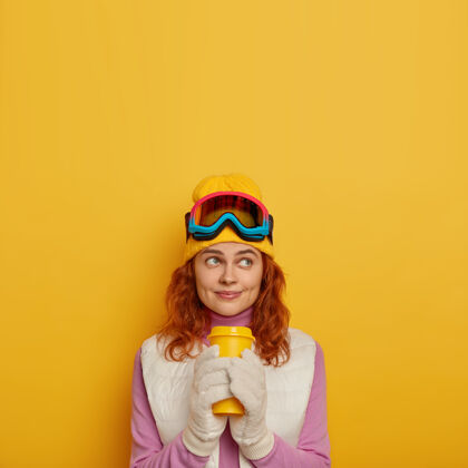 红发梦寐以求的女人拿着一次性杯子里的热饮料 梦见冬天的假期 头戴滑雪眼镜 头戴帽子 站在室内护目镜红发装备
