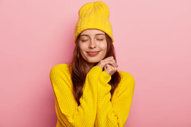 女人可爱的年轻欧洲女人的肖像闭上眼睛 穿着鲜艳的黄色针织毛衣和头饰 隔离在粉红色的背景上白种人服装请