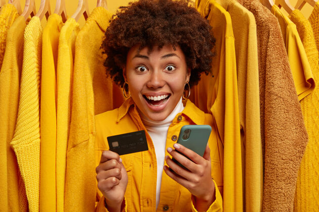 年轻情绪开朗的购物女用手机上网付款 拿着信用卡 站在衣架上的黄色毛衣之间购物狂一室内