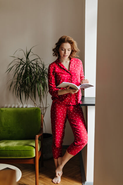 早晨赤脚女士饶有兴致地读杂志的全景客厅里穿着红色睡衣的快乐女人的室内镜头女人卷发人