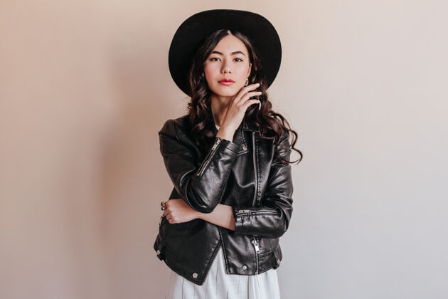 人戴着帽子沉思的亚洲年轻女子看着镜头穿着皮夹克的日本女子站在米色背景上生活方式休闲灵感